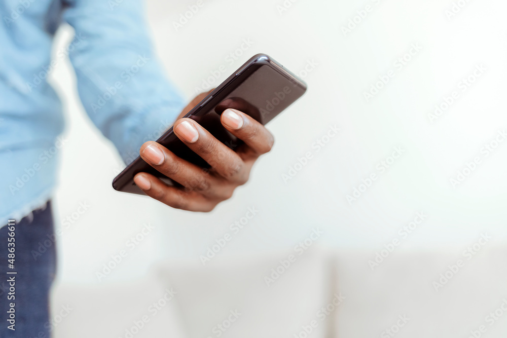 Handsome young African man looking on phone and smiling while sitting on the couch at home. Young happy black man at home sending messages on smartphone lying on beige couch in living room, copy space