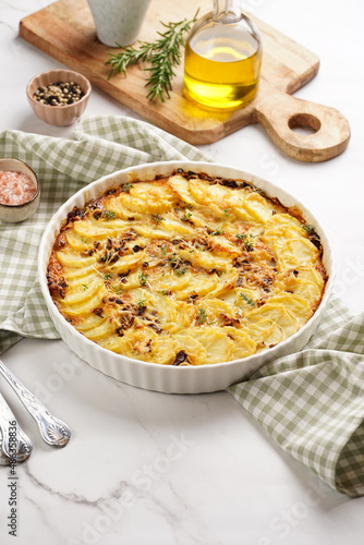 Slices potato casserole with red onions, fresh thyme and rosemary on green checkered kitchen towel , wooden board with flavoring ingredients: pepper, salt, olive oil