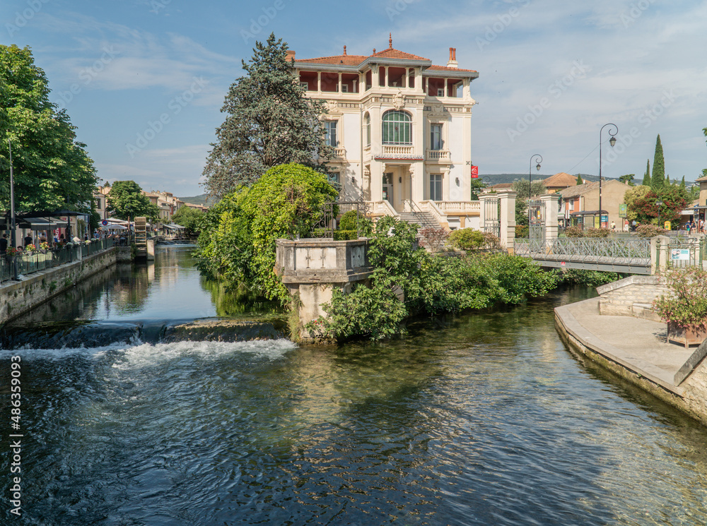 L'isle-sur-la-Sorgue, Vaucluse - France - July 2 2021: The river Sorgue divided in two crossing in the middle of a French village with views of a small bridge and a big white House.