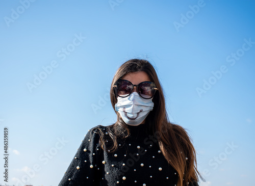 mujer caucásica con gafas de sol y cabello castaño con una mascarilla dibujada una sonrisa en un bonito día soleado.