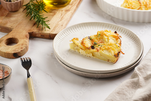 Slice of potato casserole with red onions, fresh thyme and rosemary on green checkered kitchen towel , wooden board with flavoring ingredients: pepper, salt, olive oil