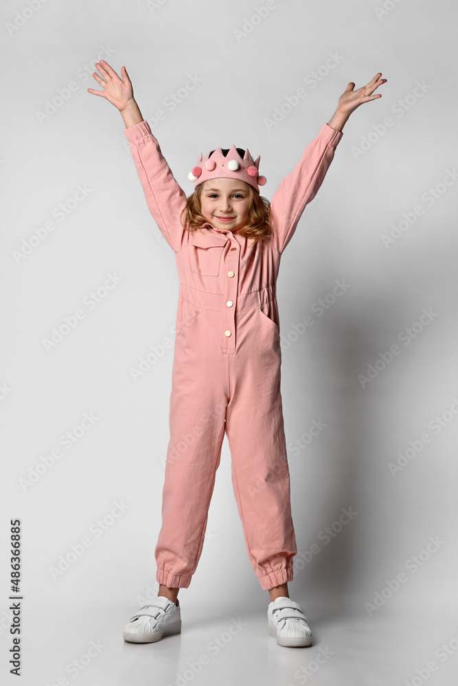 cute girl with a plush crown, stands in full growth with her hands up. in a cotton summer jumpsuit, dusty rose color, with comfortable pockets. on a light background. Children's fashion. cute girl with a plush crown, stands in full growth with her hands up. in a cotton summer jumpsuit, dusty rose color, with comfortable pockets. on a light background. Children's fashion.