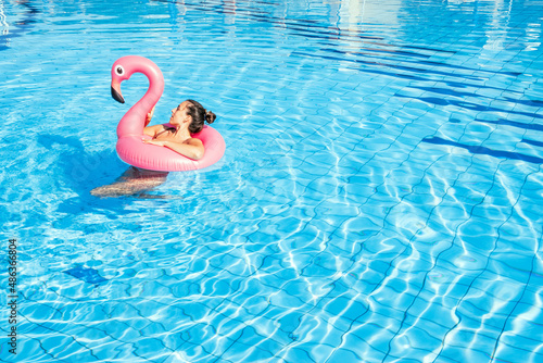 Female by pool. Happy young sexy girl in bikini swimsuit with pink flamingo float in blue water. Luxury lifestyle travel.