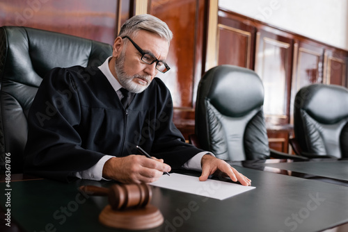 Photo senior judge in robe and eyeglasses holding pen near paper and blurred gavel