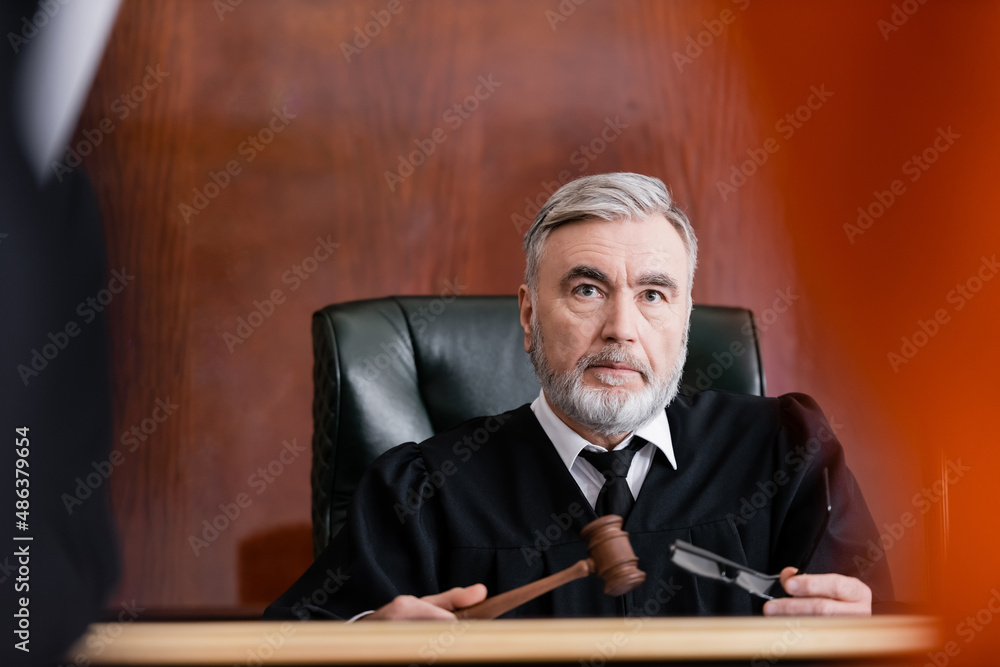 serious judge holding gavel and eyeglasses in court on blurred foreground serious judge holding gavel and eyeglasses in court on blurred foreground