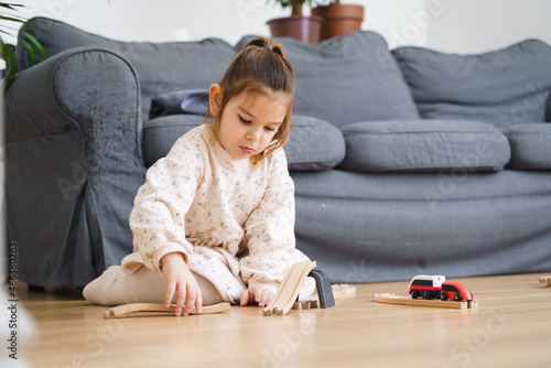 Toddler girl in white dress plays with wooden train at home in the living room 
