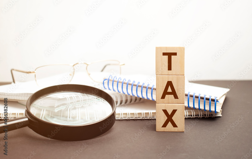 the word TAX is written on wooden cubes on a gray background. close-up of wooden elements, magnifying glass, paper documents and coins on a notebook