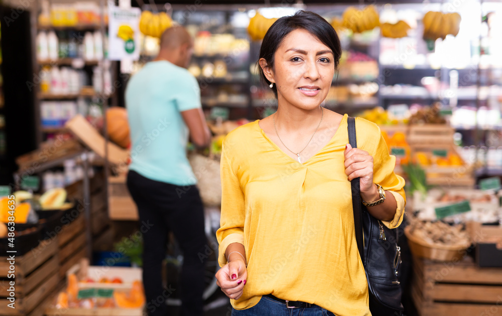 Portrait of smiling Hispanic woman choosing food products on shelves in grocery shop
