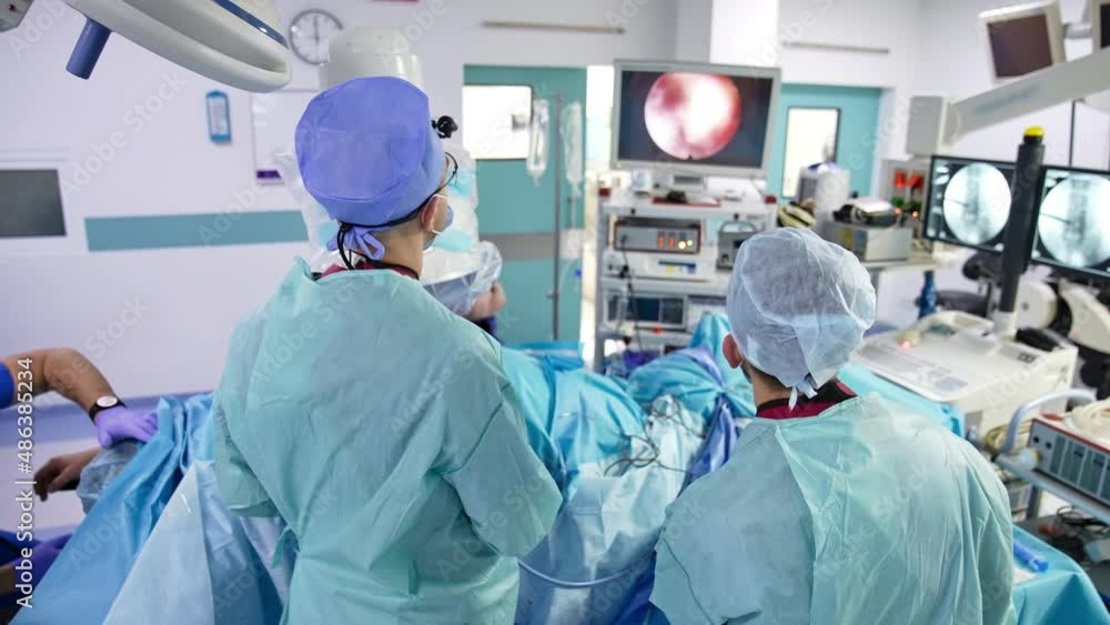 Doctors standing with their backs to the camera over the patient. Surgeon and assistant look at the screen showing operated organ. Well-equipped surgery room background. Doctors standing with their backs to the camera over the patient. Surgeon and assistant look at the screen showing operated organ. Well-equipped surgery room background.