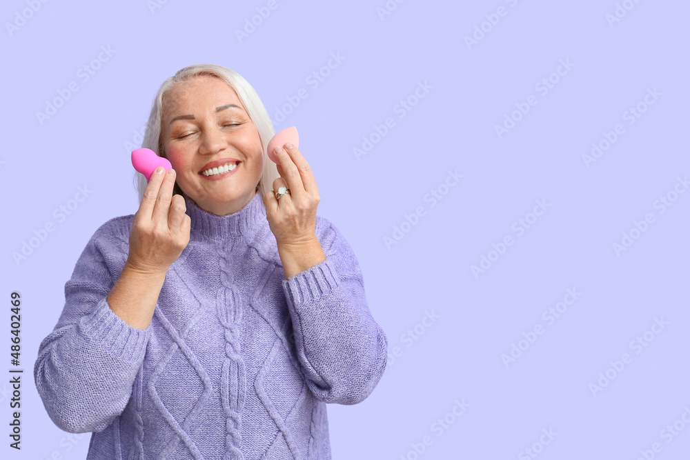 Mature woman in knitted sweater holding beauty blenders on violet background. International Women's Day celebration Mature woman in knitted sweater holding beauty blenders on violet background. International Women's Day celebration