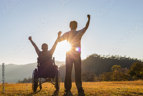 Young girl sitting in wheelchair raised hands with his care helper in sunset mountain park. Silhouette