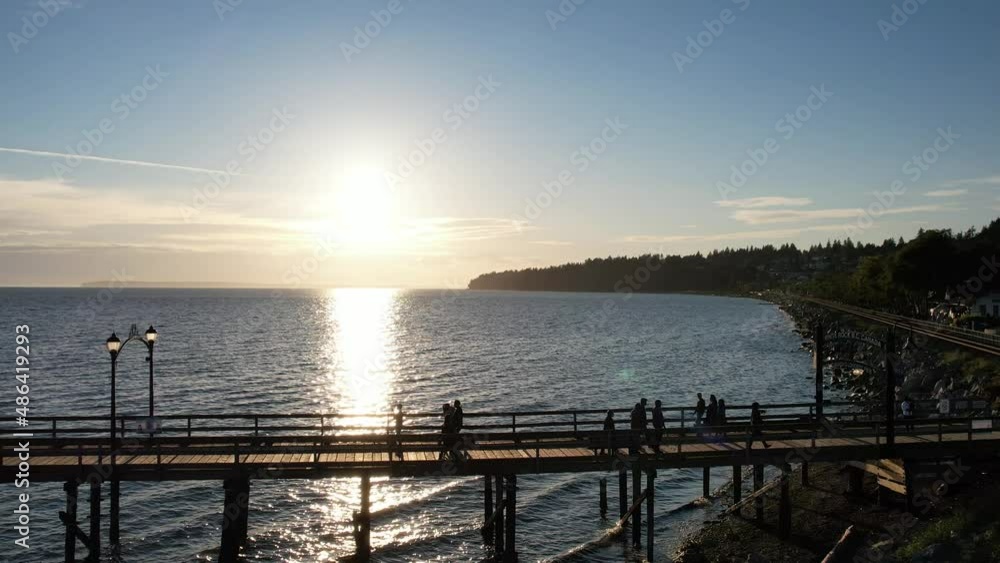 Silhouettes of people walking on a wooden pier with the sun against the lens. 4K aerial drone video of people having a leisure activity. Somewhere along the Canadian coast. Silhouettes of people walking on a wooden pier with the sun against the lens. 4K aerial drone video of people having a leisure activity. Somewhere along the Canadian coast.