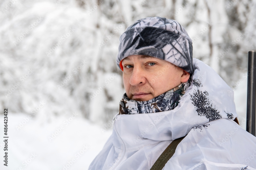 Obraz premium Portrait of a male hunter in a winter forest with a gun in a camouflage white suit. The concept of winter hunting.