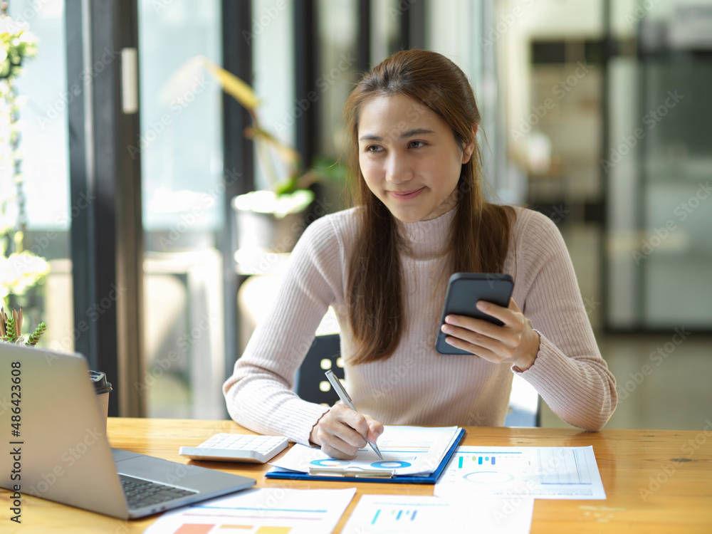Female worker holding a smartphone while working on her paperwork Female worker holding a smartphone while working on her paperwork
