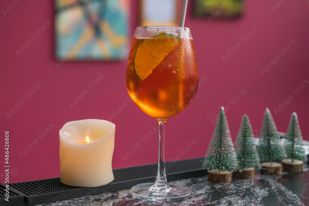 Aperol spritz summer cocktail in mister glass with lime, eco friendly straw and bubbles. Close up. Pink background. Toy Christmas trees, candle. Aperol spritz summer cocktail in mister glass with lime, eco friendly straw and bubbles. Close up. Pink background. Toy Christmas trees, candle.