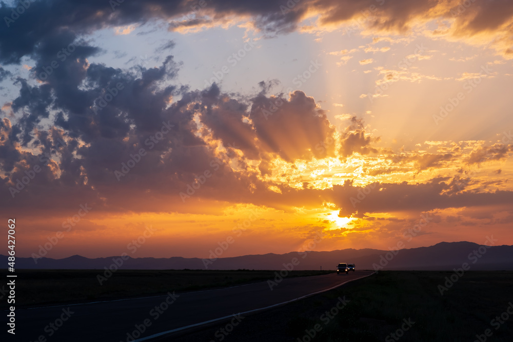Beautiful sunset with cloudy sky and sun rays. Sunrise with road, mountains silhouettes and orange golden clouds.