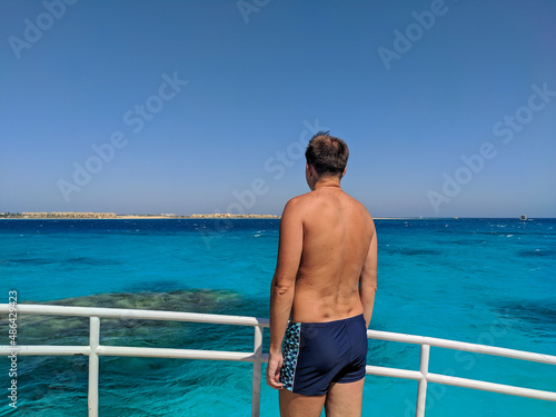 Hurghada, Egypt - October 1, 2021: Man in swimming trunks stands on board the yacht and looks at the azure red sea. Copy space.
