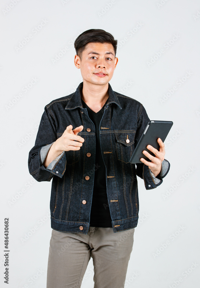 Portrait studio shot Asian young confident handsome cool short hair male hipster model wearing casual street denim jeans jacket standing holding tablet computer pointing at camera on gray background Portrait studio shot Asian young confident handsome cool short hair male hipster model wearing casual street denim jeans jacket standing holding tablet computer pointing at camera on gray background