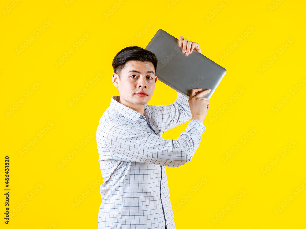 Portrait studio shot of millennial Asian young male businessman in casual outfit standing smiling holding laptop computer on yellow background Portrait studio shot of millennial Asian young male businessman in casual outfit standing smiling holding laptop computer on yellow background