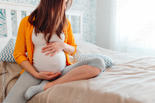 Pregnant woman holds hands on belly in bed on bright background.