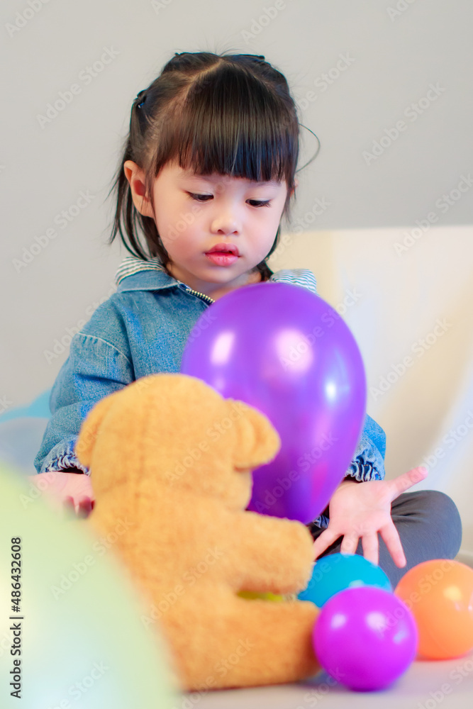 Studio shot of little cute preschooler girl daughter in casual street wears jeans jacket and sneakers sitting smiling teddy bear doll on floor playing with colorful plastic balls on gray background Studio shot of little cute preschooler girl daughter in casual street wears jeans jacket and sneakers sitting smiling teddy bear doll on floor playing with colorful plastic balls on gray background