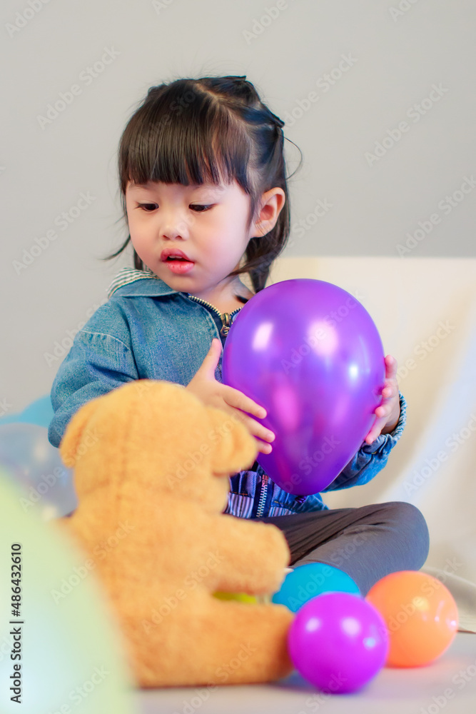 Studio shot of little cute preschooler girl daughter in casual street wears jeans jacket and sneakers sitting smiling teddy bear doll on floor playing with colorful plastic balls on gray background Studio shot of little cute preschooler girl daughter in casual street wears jeans jacket and sneakers sitting smiling teddy bear doll on floor playing with colorful plastic balls on gray background
