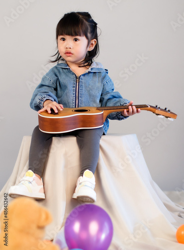 Studio shot of little cute preschooler girl daughter in jeans jacket sitting on stand holding small guitar ukulele learning playing classical music instrument lesson in classroom on gray background