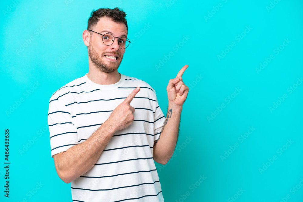 Young Brazilian man isolated on blue background frightened and pointing to the side Young Brazilian man isolated on blue background frightened and pointing to the side