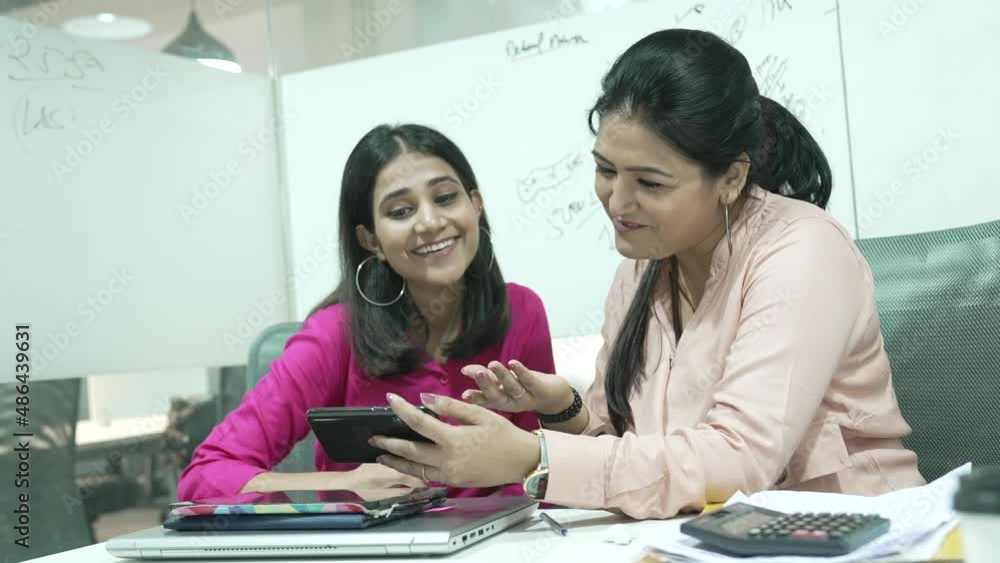 young Indian two woman are laughing while watching a funny video on a smartphone or cellphone sitting at office room.Two happy friends watching a movie on a mobile phone. young Indian two woman are laughing while watching a funny video on a smartphone or cellphone sitting at office room.Two happy friends watching a movie on a mobile phone.