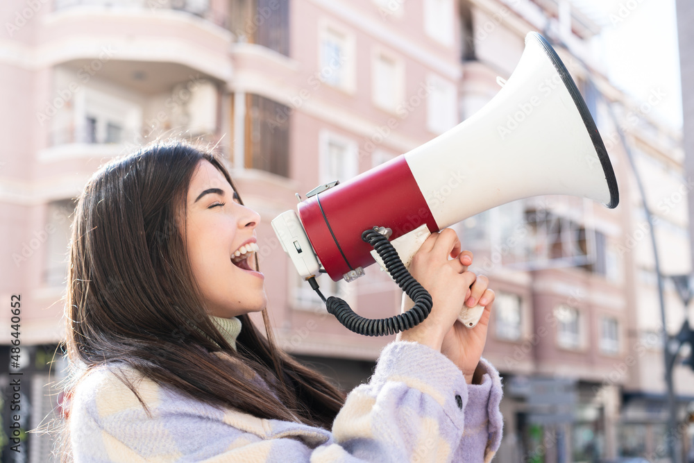 Young Brazilian woman at outdoors shouting through a megaphone to announce something in lateral position - obrazy, fototapety, plakaty 
