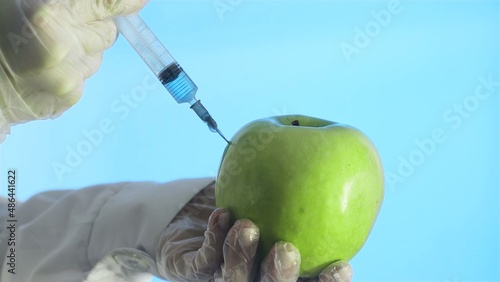 Close-up of a human in a medical coat and gloves injecting a syringe into an apple with some liquid on a blue background