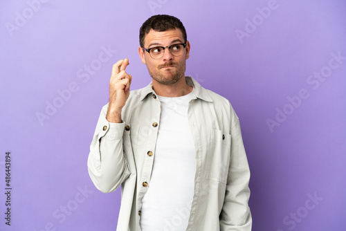Brazilian man over isolated purple background with fingers crossing and wishing the best
