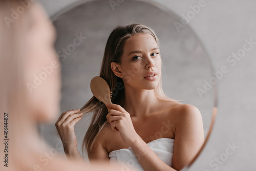Young cute lady wrapped in towel, standing in front of mirror at bathroom and combing her hair after shower