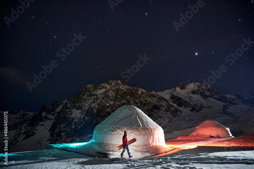 A man with a snowboard walking near a yurt at night in winter in the mountains photo