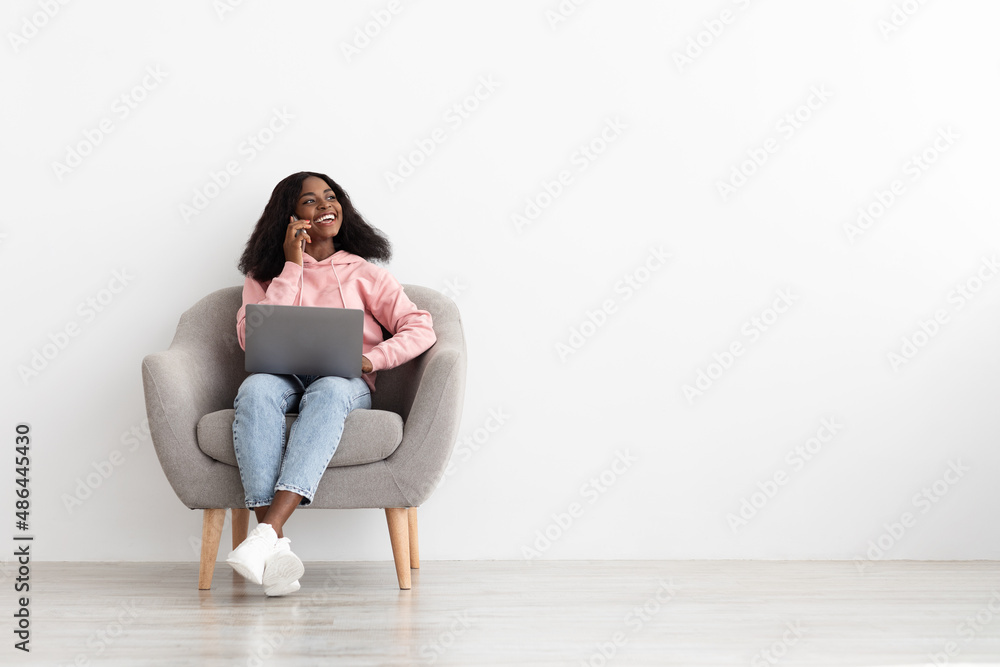 Happy young black woman having phone conversation, using laptop