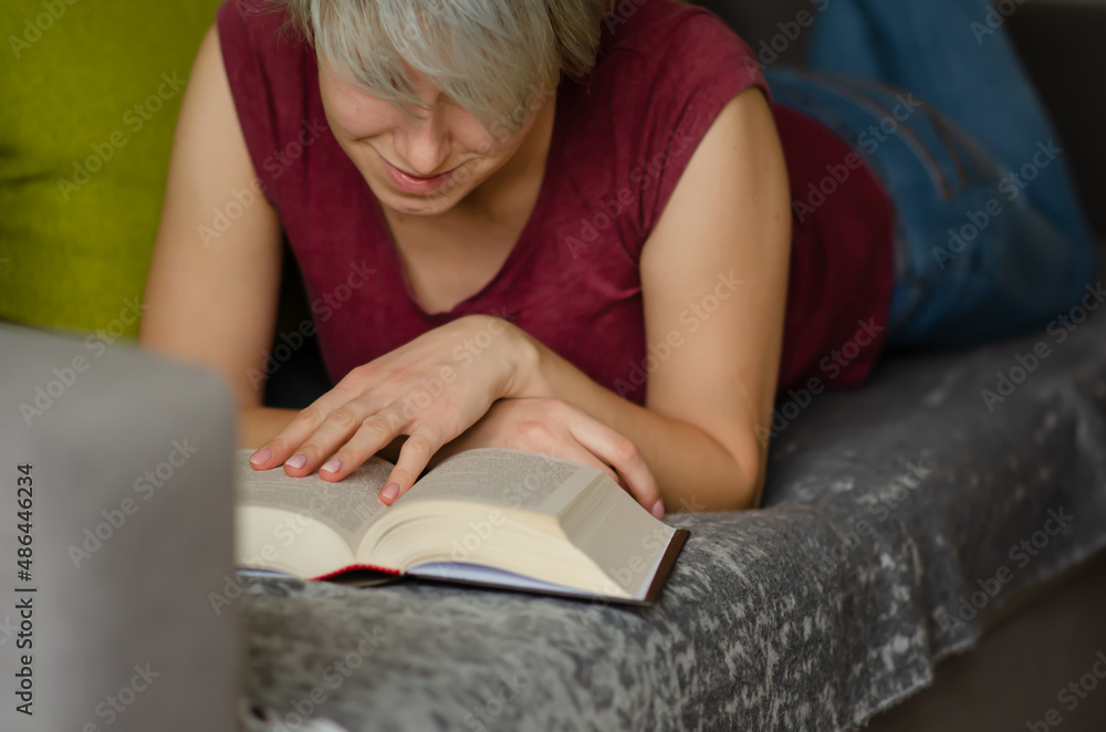 Selective focus on book read by caucasian woman with short haircut in burgundy T-shirt and jeans lying on gray sofa with pillows. Young woman reads an interesting book and laughs.