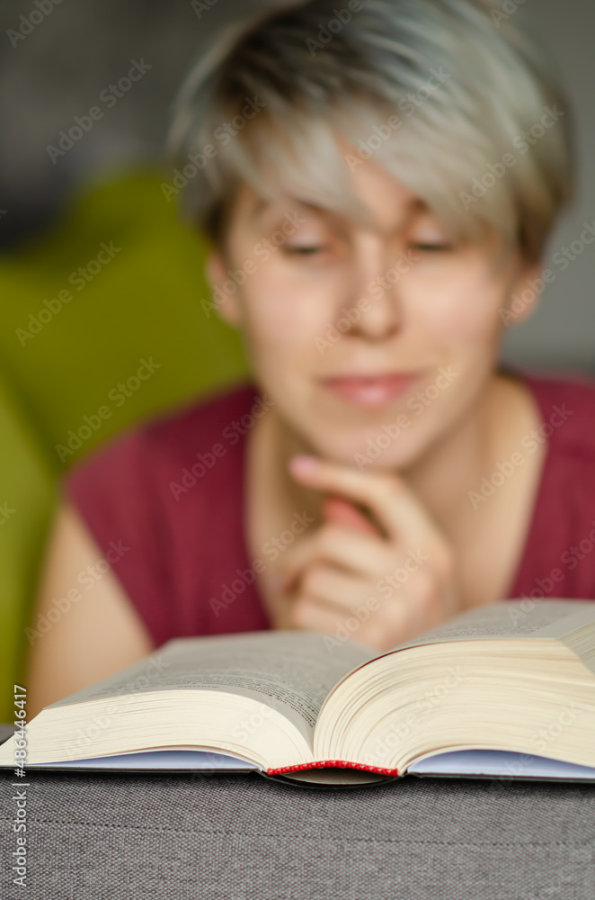 Selective focus on book read by smiling caucasian woman with short haircut on gray sofa. Concept of time alone with yourself. Young woman reads an interesting book and smiling. Selective focus on book read by smiling caucasian woman with short haircut on gray sofa. Concept of time alone with yourself. Young woman reads an interesting book and smiling.