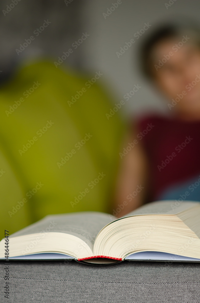 Selective focus on book lies on gray sofa on blurred background of caucasian woman in burgundy T-shirt and jeans lying on sofa with pillows. Concept of time for reading. Selective focus on book lies on gray sofa on blurred background of caucasian woman in burgundy T-shirt and jeans lying on sofa with pillows. Concept of time for reading.