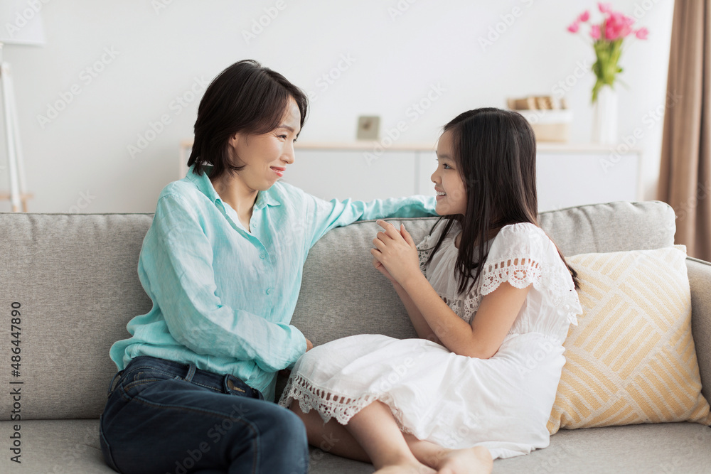 Happy little Asian girl speaking to her smiling grandmother, sitting on couch together at home Happy little Asian girl speaking to her smiling grandmother, sitting on couch together at home