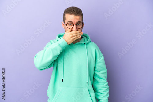Young handsome caucasian man isolated on purple background covering mouth with hand © luismolinero