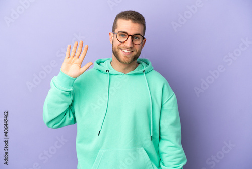 Young handsome caucasian man isolated on purple background saluting with hand with happy expression © luismolinero