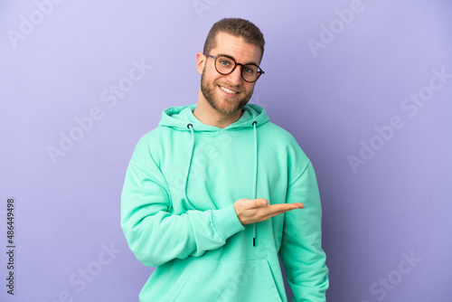 Young handsome caucasian man isolated on purple background presenting an idea while looking smiling towards