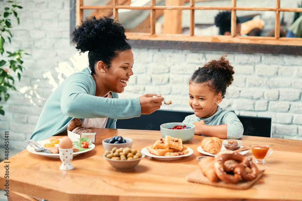 Happy black mother feeds her small daughter during breakfast at dining table.