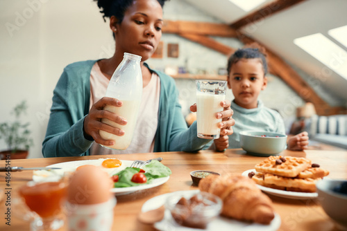 Close-up of black mother pours milk into daughter's glass while having breakfast together at dining table.