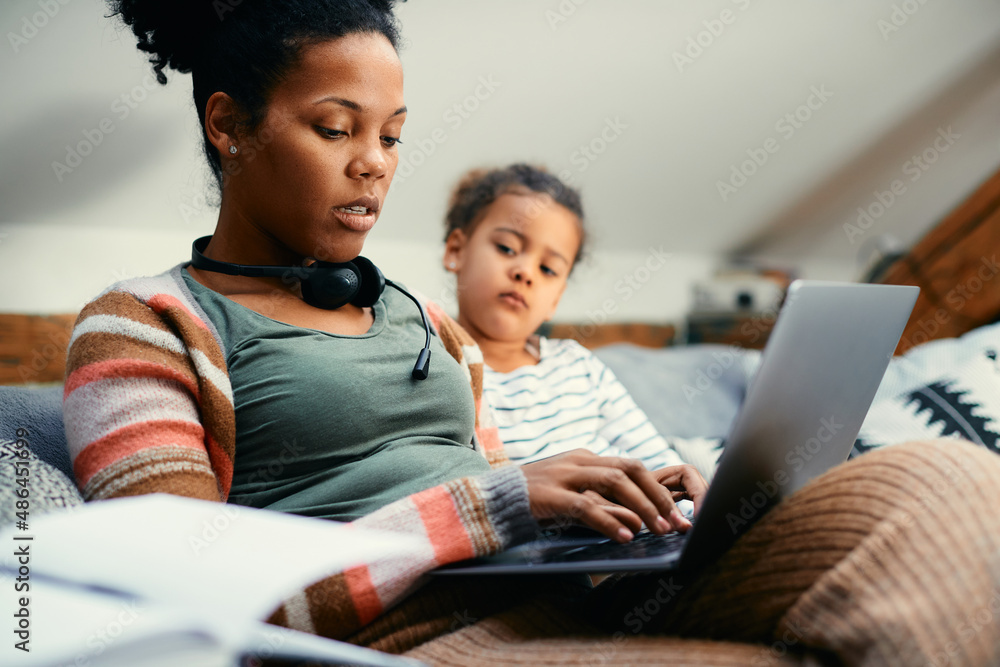 African American mother works on laptop while  while being with her daughter at home.