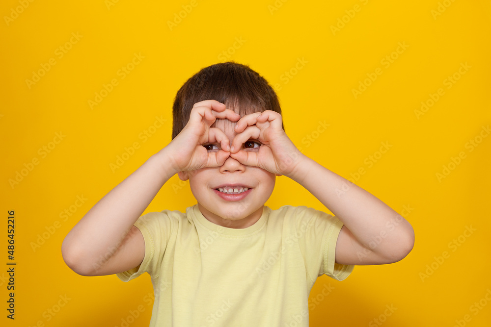 Portrait of a happy boy holding his hands in the form of glasses in front of eyes and laughing . Positive emotions, playful child smiles, childhood, school age. Yelow background. Copy space. Portrait of a happy boy holding his hands in the form of glasses in front of eyes and laughing . Positive emotions, playful child smiles, childhood, school age. Yelow background. Copy space.