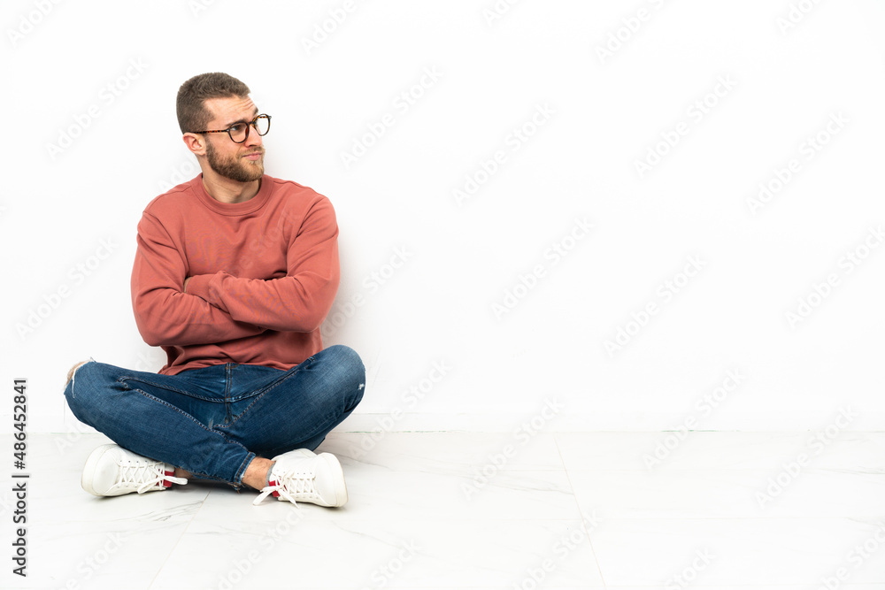 Young handsome man sitting on the floor making doubts gesture while lifting the shoulders Young handsome man sitting on the floor making doubts gesture while lifting the shoulders