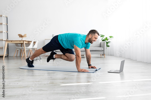 Fototapeta Naklejka Na Ścianę i Meble -  Happy active young european man doing leg exercises on mat on floor and looking at laptop in living room