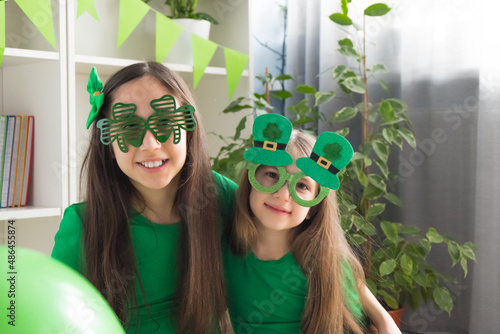 Two sister girls in funny green glasses, dressed in green t-shirts, hold balloons and have fun at home celebrating St. Patrick's Day.