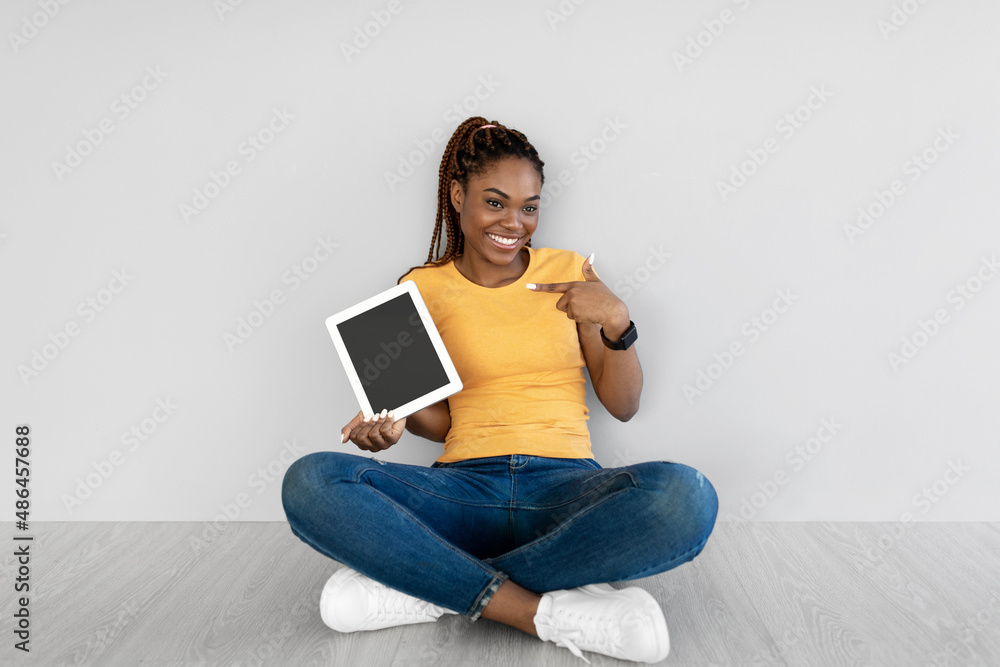 Black woman pointing at tablet with blank screen, sitting on floor against grey studio wall, mockup for website or app Black woman pointing at tablet with blank screen, sitting on floor against grey studio wall, mockup for website or app